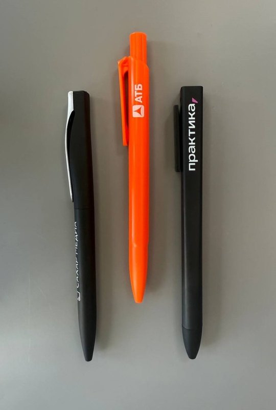 Три ручки с логотипами на сером фоне: две чёрные и одна оранжевая.